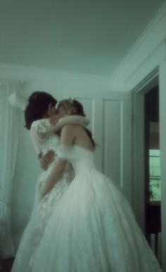 two women in wedding gowns hugging each other while standing next to a doorway with white curtains