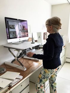 a woman standing in front of a desk with a computer on it and looking at the screen