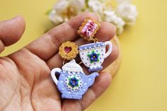 a hand holding three miniature teapots with hearts on them and flowers in the background