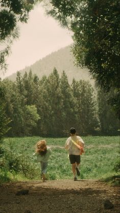 two people walking down a dirt road in front of green grass and trees with mountains in the background