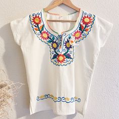 a white blouse with colorful flowers on it hanging from a wooden hanger against a wall