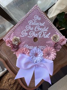 a pink graduation cap decorated with flowers and ribbon on top of a wooden chair in front of a window