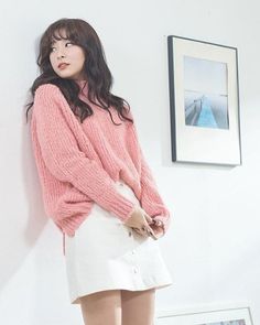 a woman standing in front of a wall wearing a pink sweater and white miniskirt