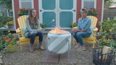 two people sitting around a fire pit in front of a red and white house with potted plants