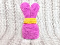 a crocheted purple bunny sitting on top of a white blanket with a yellow ribbon around its neck