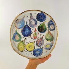 a hand holding a plate with different colored fruit designs on the front and sides, in gold trim