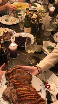 people are sitting at a table with food and wine in front of them, one person is holding a plate that has meat on it