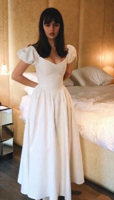 a woman standing in front of a bed wearing a white dress with ruffle sleeves