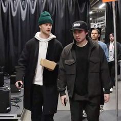 two young men are walking down the street in front of a black backdrop and one is wearing a green beanie