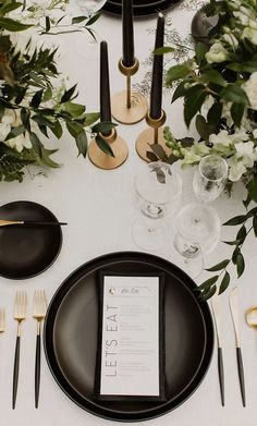 the table is set with black plates and silverware, gold flatware, greenery, and white flowers