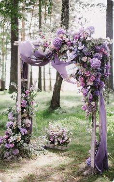 an outdoor wedding ceremony with purple flowers and greenery on the arbor, surrounded by tall trees