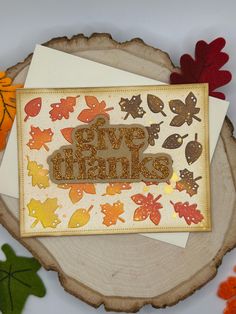 a card with the words give thanks surrounded by fall leaves and acorns on a tree stump