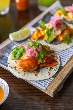 two tacos with meat, vegetables and sauces on a wooden tray next to drinks