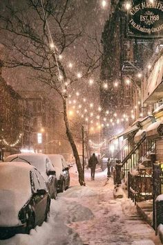 a city street covered in snow at night with people walking on the sidewalk and parked cars