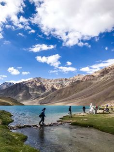 people are standing on the shore of a body of water with mountains in the background