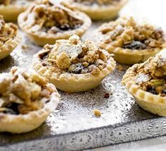 many small pies are sitting on a silver tray with powdered sugar and crumbled toppings