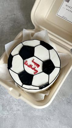 a cake in the shape of a soccer ball with red writing on it, sitting inside an open plastic container