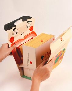 a person is holding an open book in front of a cardboard box with a face on it