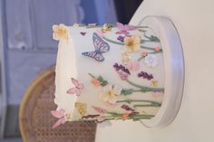 a white cake with flowers and butterflies on it sitting on a table next to a wicker chair