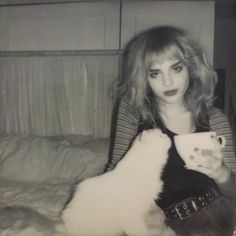 a woman sitting on a bed holding a white and black cat in her lap while drinking from a cup