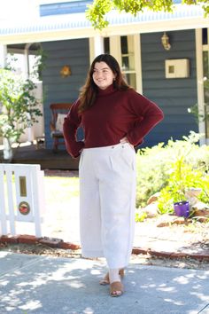 a woman standing in front of a house wearing white pants and a red sweater with her hands on her hips