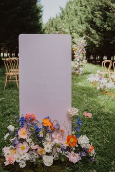 a sign that is sitting in the grass with flowers around it and some chairs behind it