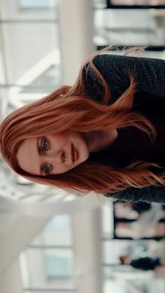 a woman with long red hair standing in front of a window looking at the camera