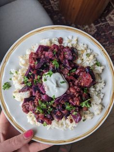 a white plate topped with rice and meat covered in sauce next to a woman's hand