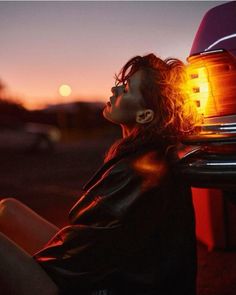 a woman sitting in front of a car with her head turned to the side and lights on