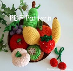 crocheted fruits and vegetables in a bowl on a white surface with greenery