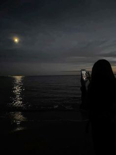 a woman standing on the beach taking a photo with her cell phone at night time