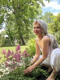 a woman kneeling down in the grass next to some plants and flowers with trees behind her