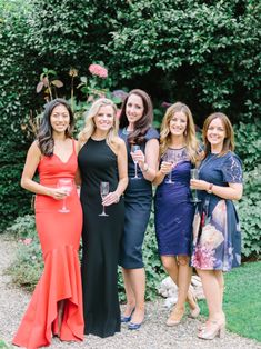 four women in dresses holding wine glasses and posing for the camera with trees behind them