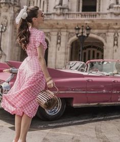 a woman standing in front of an old pink car with a bow in her hair