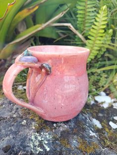 a pink coffee cup sitting on top of a rock next to green plants and moss