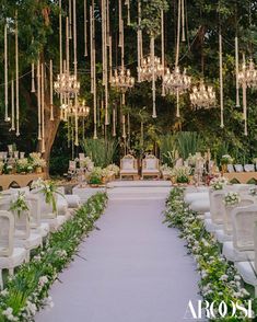 an outdoor wedding setup with white chairs and chandeliers hanging from the ceiling, surrounded by greenery