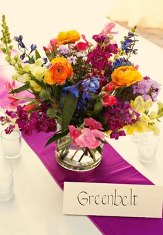 a vase filled with lots of colorful flowers on top of a purple table runner next to two glasses