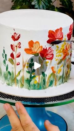 a hand is pointing at a decorated cake on a table with flowers painted on it