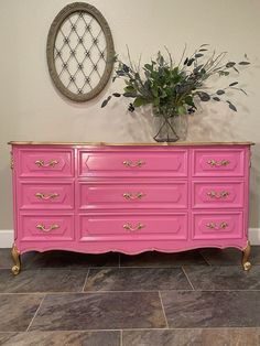 a pink dresser with gold trim and flowers in a vase next to it on a tile floor