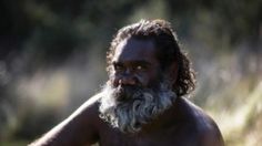 a man with white beard and no shirt sitting on the ground in front of trees