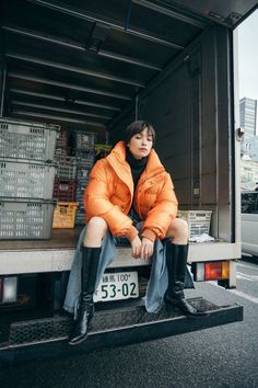 a woman in an orange jacket sitting on the back of a truck with her legs crossed