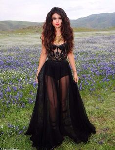 a woman in a black dress standing in a field with purple flowers and mountains behind her