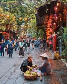 two women sitting on the ground with baskets full of fruit in front of them and people walking down the street