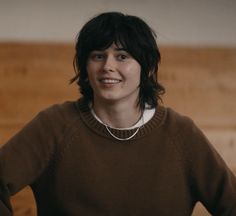 a woman standing in front of a wooden wall wearing a brown sweater and white necklace