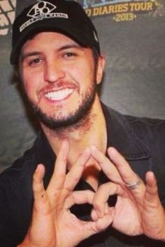 a smiling man making the v sign with his hands while wearing a black baseball cap