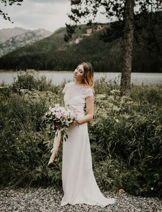 a woman in a white dress holding a bouquet standing next to some bushes and trees