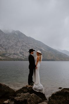 a bride and groom standing on rocks by the water with mountains in the back ground