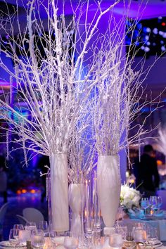 two vases filled with white branches and candles on a table in front of a blue lit room