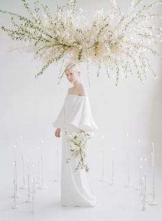 a woman in a white dress is standing next to candles and flowers with her hands on her hips