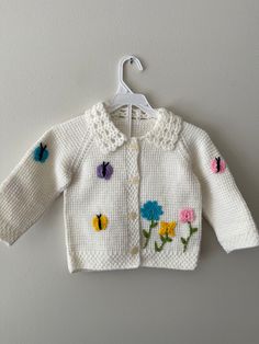 a white knitted sweater with flowers and butterflies on it, hanging from a hanger